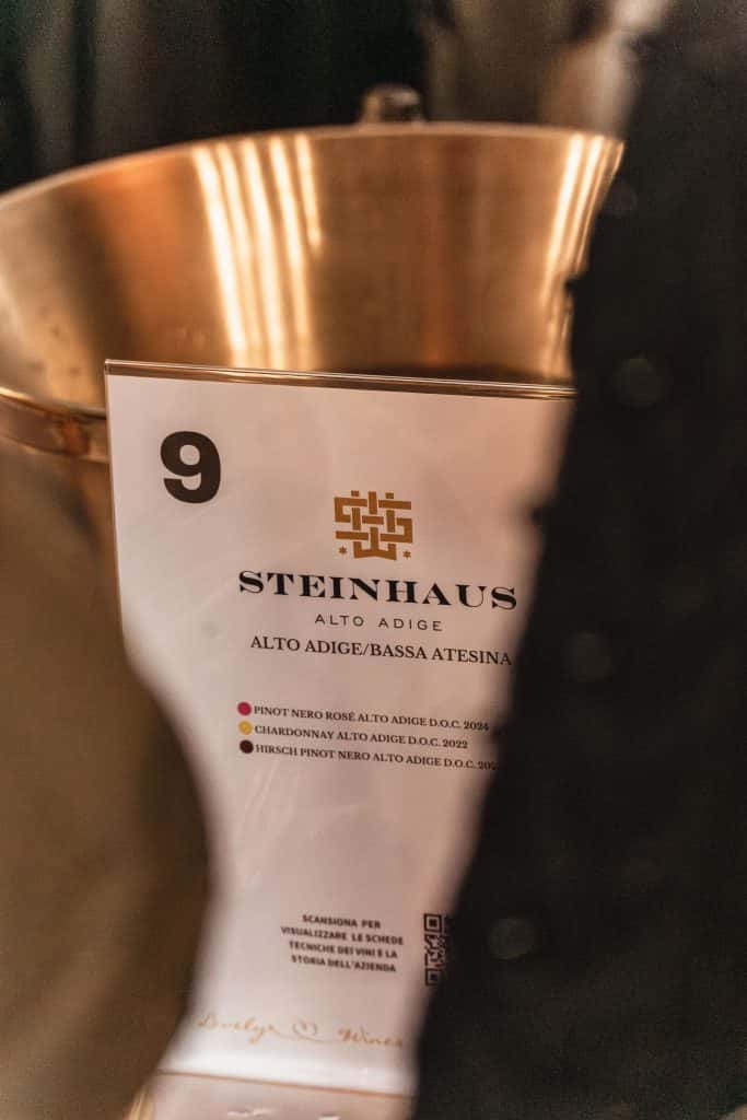 Primo piano di una scheda informativa sul vino Steinhaus Alto Adige con il numero 9, che elenca diversi tipi di vino, posta davanti a un contenitore in ottone. Oggetti parzialmente oscurati e primo piano sfocato circondano la scheda.