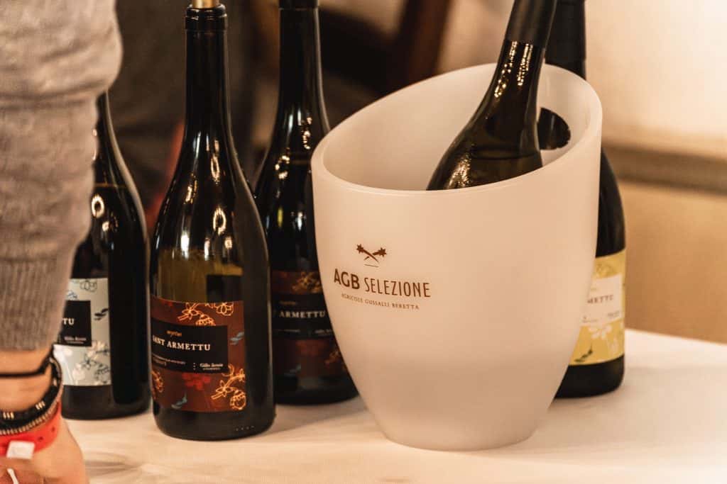 Primo piano di diverse bottiglie di vino su un tavolo, con una bottiglia che si raffredda in un secchiello per il ghiaccio bianco smerigliato con la scritta AGB SELEZIONE. Il braccio di una persona è parzialmente visibile sul lato sinistro dell'immagine.
