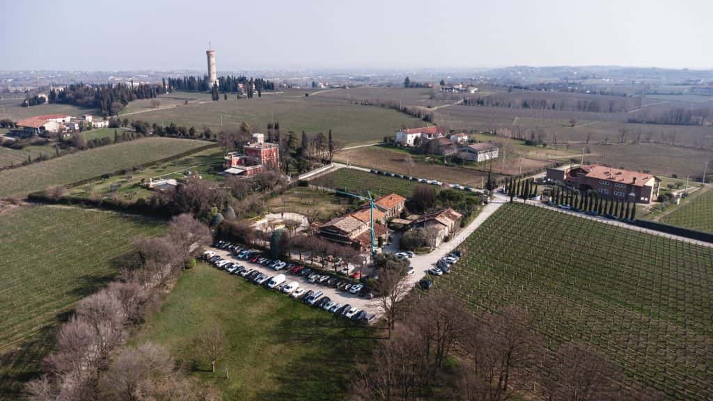 Veduta aerea di un'area rurale con vigneti, case sparse, un parcheggio pieno di auto e un'alta torre sullo sfondo; campi e alberi circondano la scena sotto un cielo nebuloso.