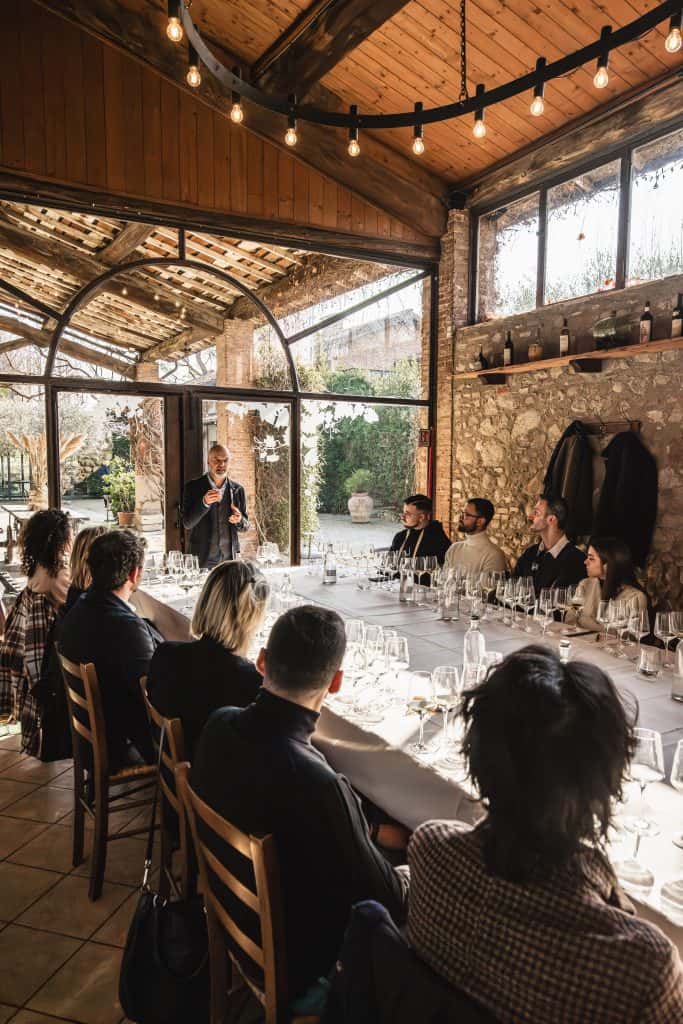 Un gruppo di persone siede intorno a un lungo tavolo con bicchieri di vino vuoti, ascoltando una persona in piedi che parla in una stanza rustica illuminata dal sole, con pareti in pietra e grandi finestre.