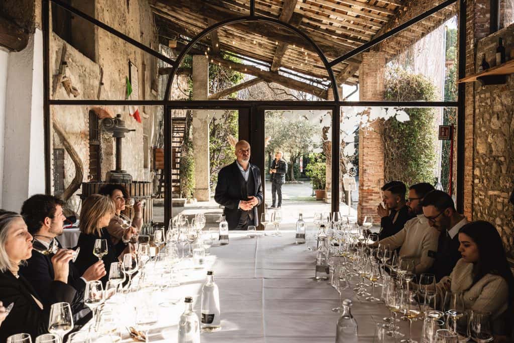 Un gruppo di persone siede intorno a un lungo tavolo apparecchiato con bicchieri di vino, mentre un uomo sta in piedi a capo, parlando, in una stanza rustica e illuminata dal sole con grandi finestre che si aprono su un giardino.