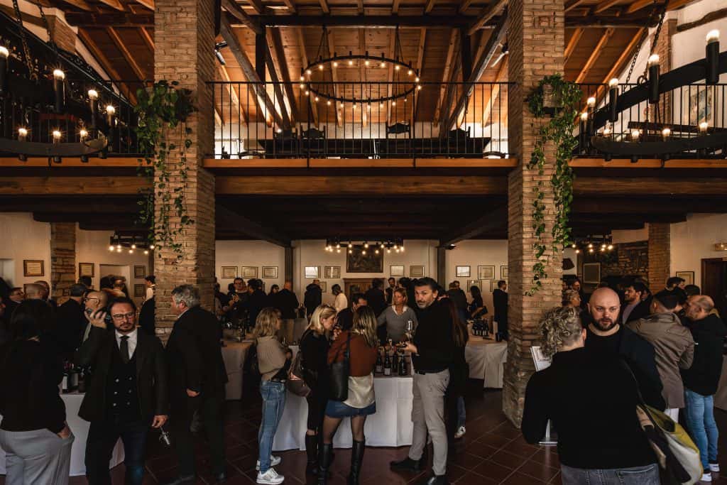 Un folto gruppo di persone si riunisce all'interno di un locale spazioso e rustico con mattoni a vista e travi in legno, socializzando e degustando vino ai tavoli coperti da teli bianchi. Le ringhiere dei balconi e le piante pendenti decorano l'interno.