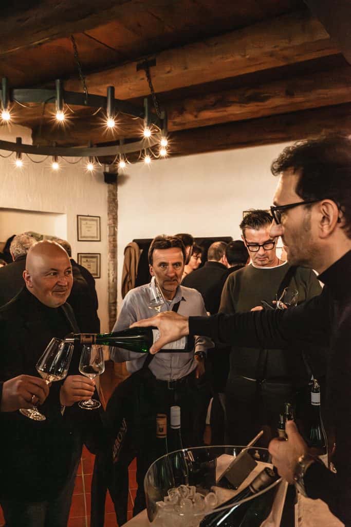 Un uomo versa del vino nei bicchieri per un gruppo di persone durante una riunione. Diversi uomini sono in piedi intorno a un tavolo con bottiglie e ghiaccio, sorridendo e tenendo in mano bicchieri di vino sotto una calda illuminazione rustica e travi di legno.