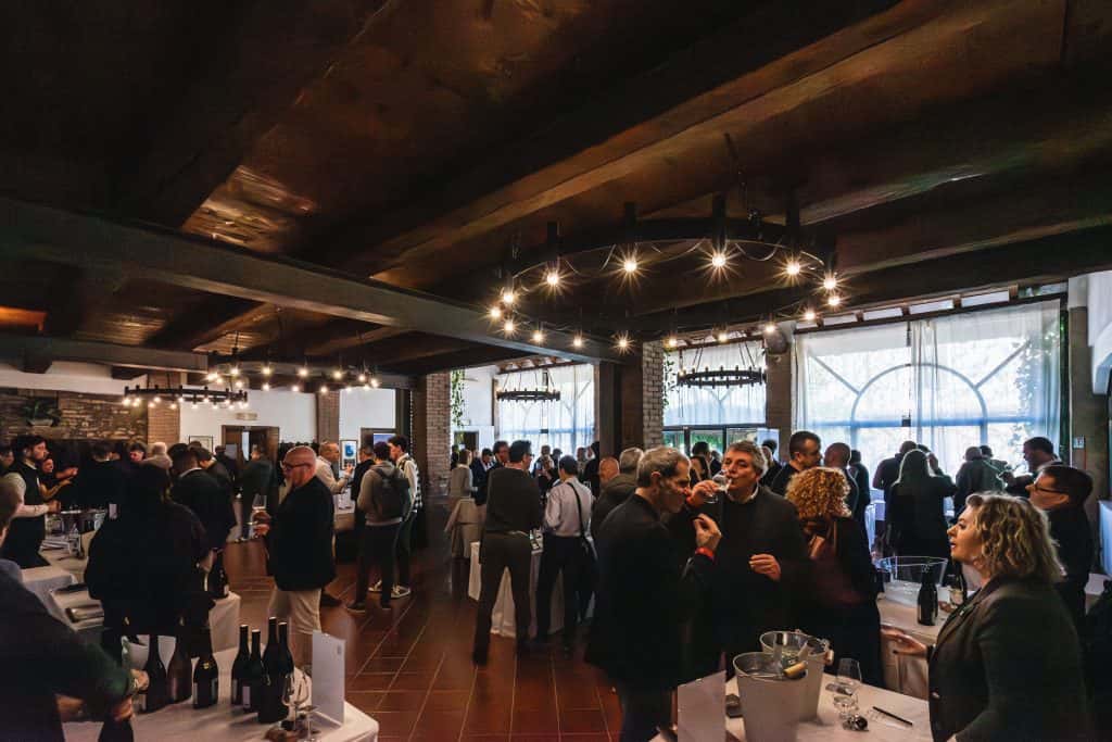 Un gruppo numeroso di persone socializza e degusta vino in un evento al chiuso con travi in legno, lampadari e grandi finestre ad arco che lasciano entrare la luce naturale. Tavoli con bottiglie e bicchieri sono disposti in tutta la stanza.
