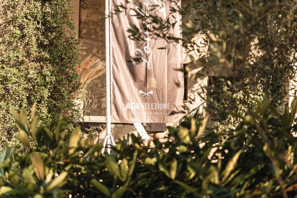 Uno striscione verticale marrone con il testo ACB SELEZIONE è esposto davanti a un muro di pietra, parzialmente nascosto da piante e alberi verdi alla luce del sole.