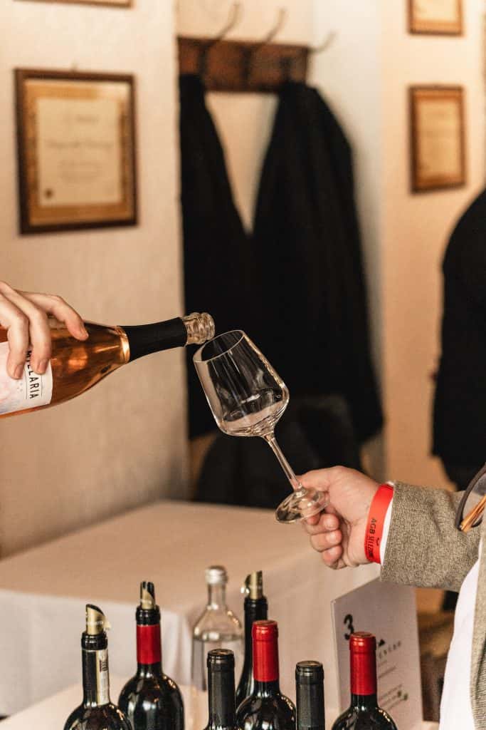 Una persona versa del vino rosé da una bottiglia in un bicchiere tenuto in mano da un'altra persona durante una degustazione di vini, con diverse bottiglie di vino e cappotti visibili sullo sfondo.