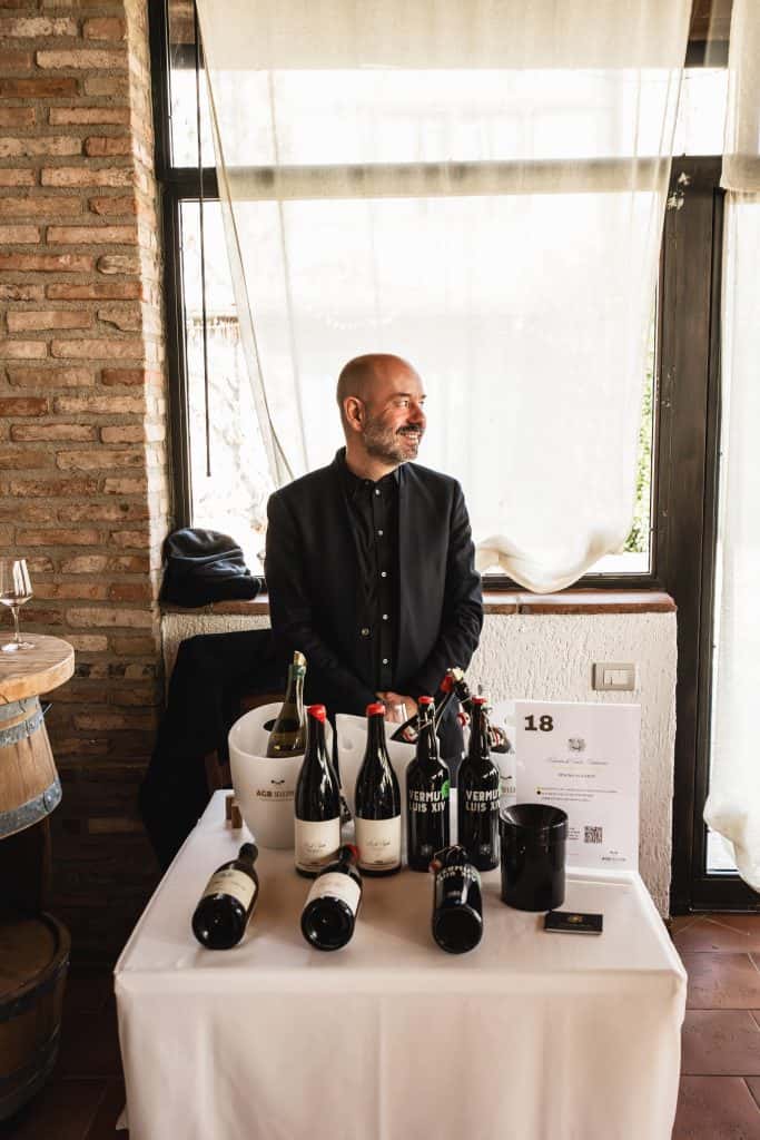 Un uomo in abito nero è in piedi dietro un tavolo che espone diverse bottiglie di vino, un dispenser e una tazza nera. Sorride e la luce naturale entra da una finestra alle sue spalle.