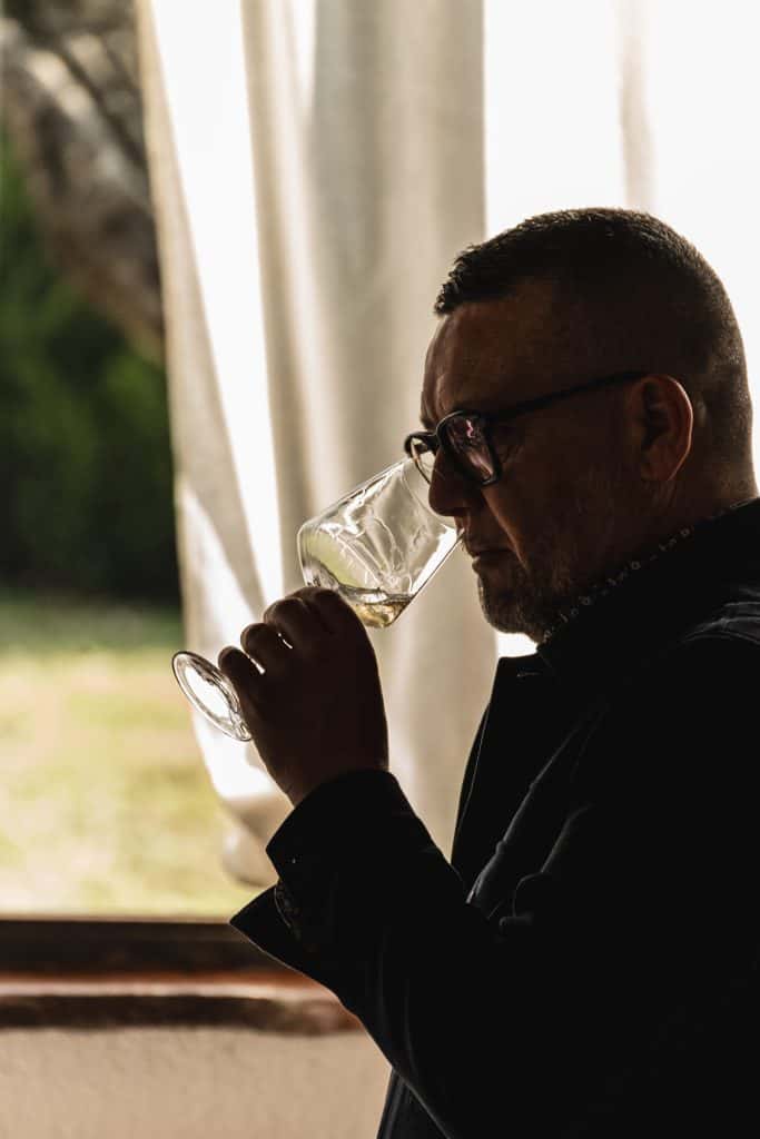 Un uomo di profilo con gli occhiali sorseggia da un bicchiere di vino, in piedi al chiuso vicino a una finestra luminosa con tende trasparenti e verde visibile all'esterno.