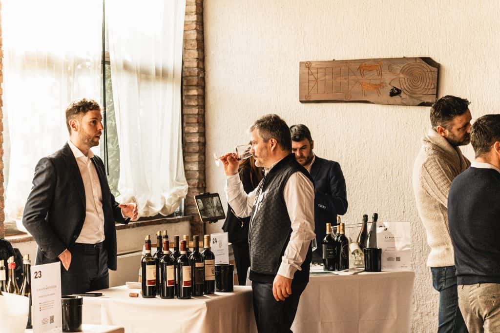 Diversi uomini sono in piedi intorno a tavoli che espongono bottiglie di vino durante un evento di degustazione. Un uomo assaggia il vino, mentre gli altri conversano. L'ambiente è illuminato da luce naturale e un muro rustico fa da sfondo.