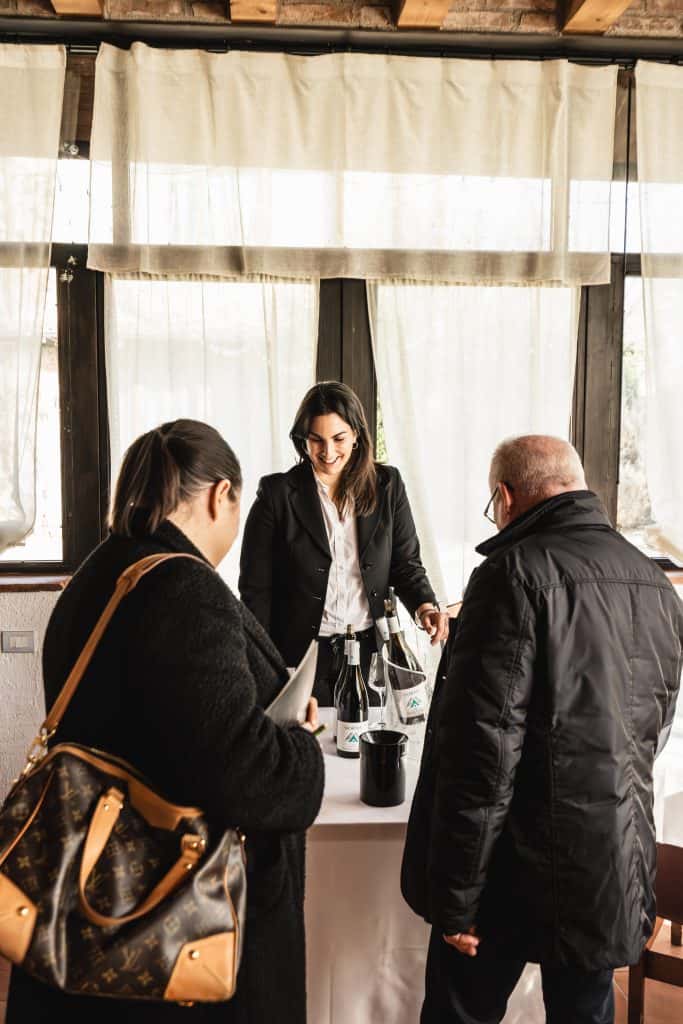 Una donna in tailleur si trova dietro un tavolo con bottiglie di vino, sorridendo e parlando con due persone in cappotto scuro che si trovano di fronte a lei in una stanza luminosa con grandi finestre.