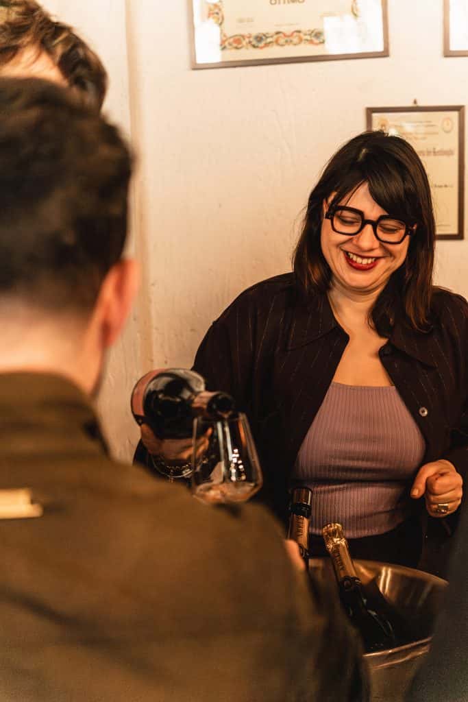 Una donna con gli occhiali sorride mentre versa del vino in un bicchiere durante un incontro sociale. È in piedi accanto a un uomo, con dei certificati appesi alla parete dietro di lei.