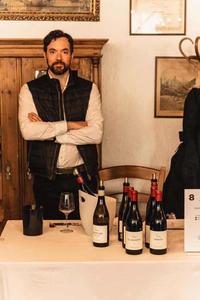 Un uomo con barba e capelli scuri è in piedi dietro un tavolo che espone diverse bottiglie di vino, un bicchiere e un secchiello per il ghiaccio. Indossa una camicia bianca e un gilet nero e tiene le braccia incrociate. L'ambiente sembra essere al chiuso.