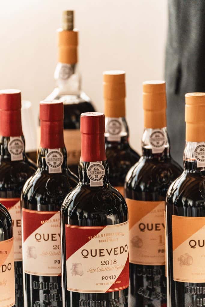 Diverse bottiglie di vino Porto Quevedo, con etichette rosse e dorate, sono disposte a stretto contatto su una superficie. Le bottiglie hanno tappi dorati e rossi e presentano etichette d'epoca.