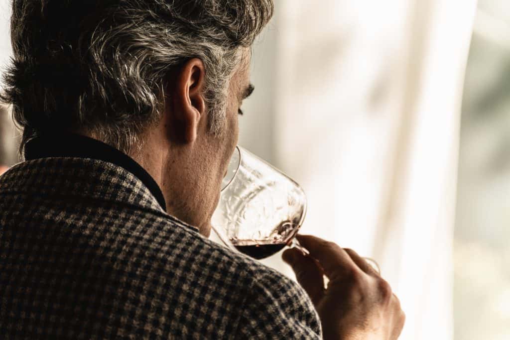 Un uomo con i capelli grigi e una giacca a scacchi tiene in mano un bicchiere di vino e annusa il vino, ripreso di spalle con una luce naturale morbida proveniente dalla finestra.