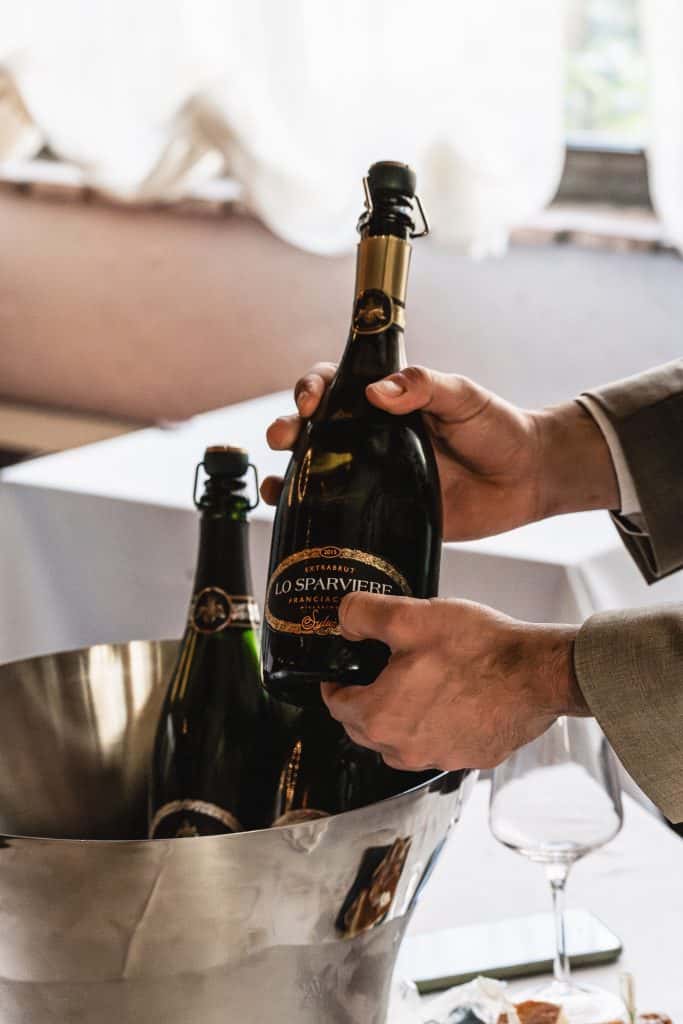 Una persona in giacca e cravatta tiene una bottiglia di spumante Lo Sparviere sopra un secchiello del ghiaccio contenente un'altra bottiglia. Un bicchiere da vino è appoggiato sul tavolo vicino. La luce filtra da una finestra sullo sfondo.