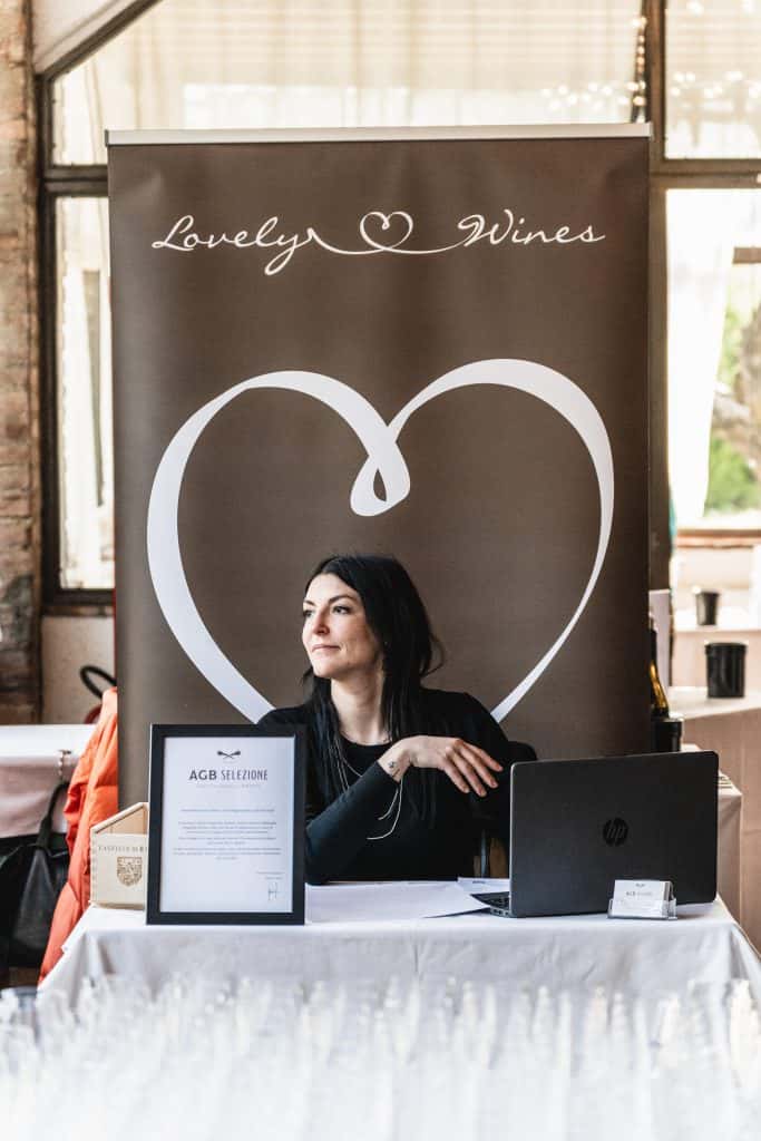 Una donna è seduta a un tavolo con un computer portatile e dei documenti, davanti a uno striscione marrone con la scritta Lovely Wines e un grande cuore sullo sfondo. Guarda di lato, lontano dalla telecamera.