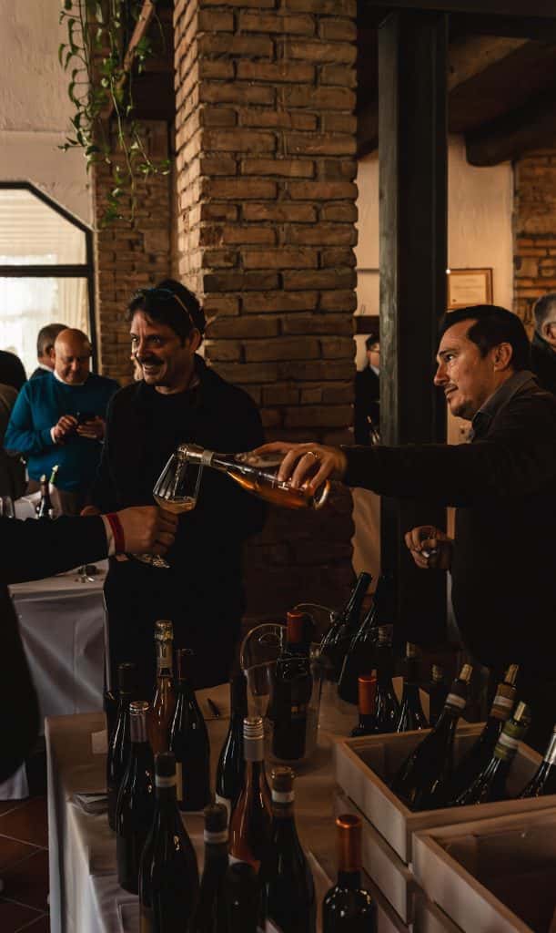 Un uomo versa il vino da una bottiglia in un bicchiere per un'altra persona durante una degustazione di vini, con bottiglie esposte sul tavolo e altre persone che si mescolano sullo sfondo.