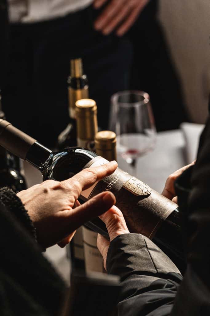 Una persona tiene in mano una bottiglia di vino, leggendone l'etichetta, con diverse altre bottiglie di vino e un bicchiere di vino rosso su un tavolo sullo sfondo. Un'altra persona appoggia la mano sul tavolo.
